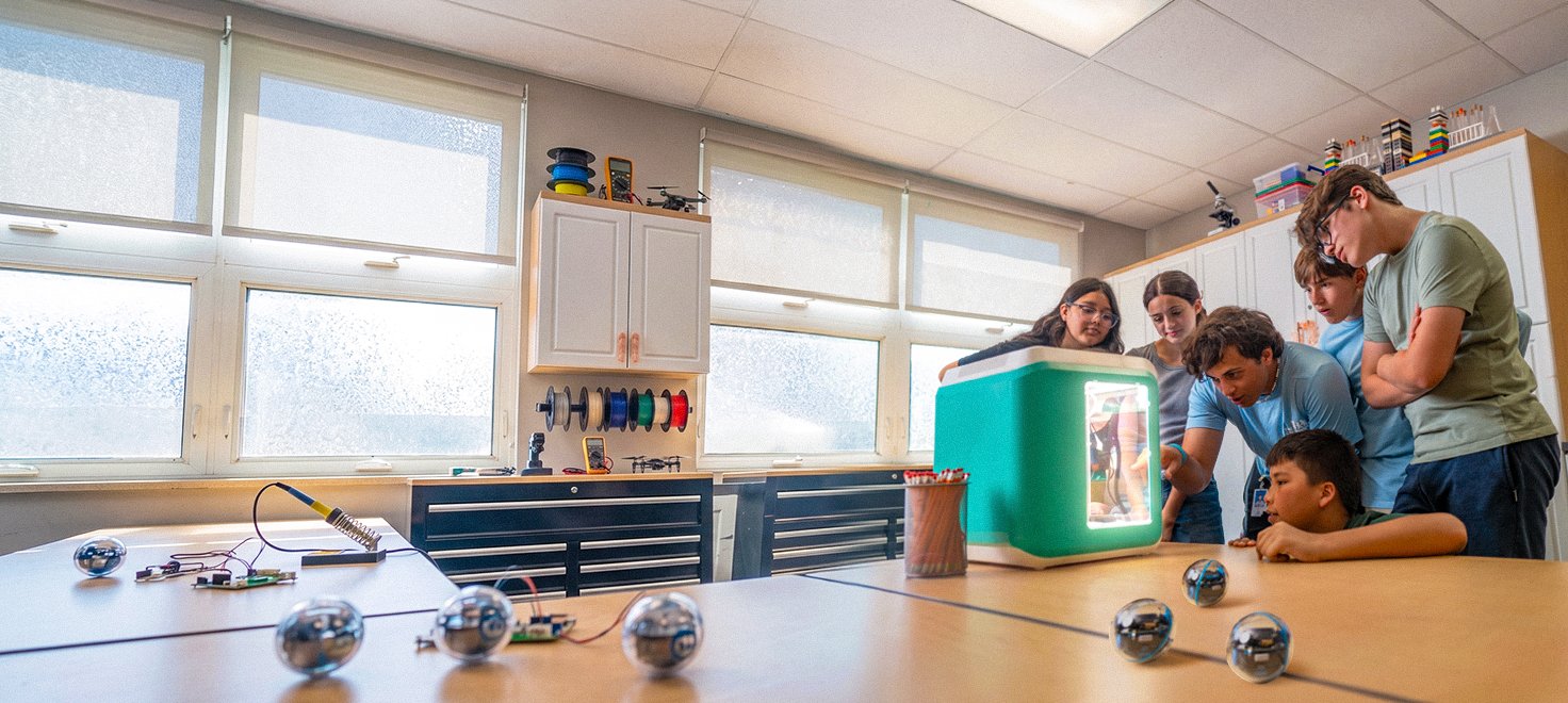 Group of teenagers huddled around a 3D printer in a classroom setting. Group of teenagers huddled around a 3D printer in a classroom setting.