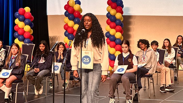 A teenage girl stands in front of a microphone on stage with other youth of similar age sitting in formation behind her.