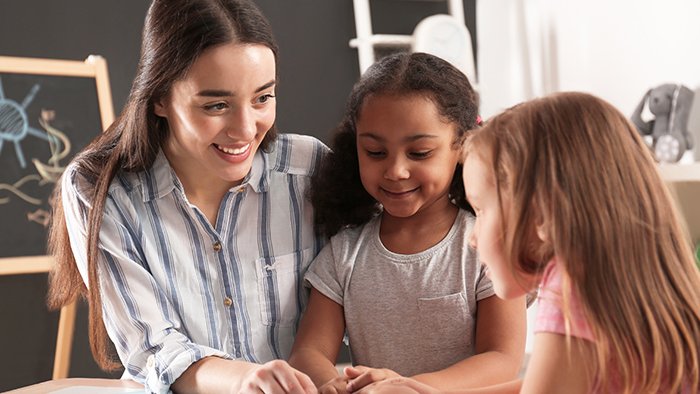 Young child care worker sits at a table reading a book to small children in kindergarten.