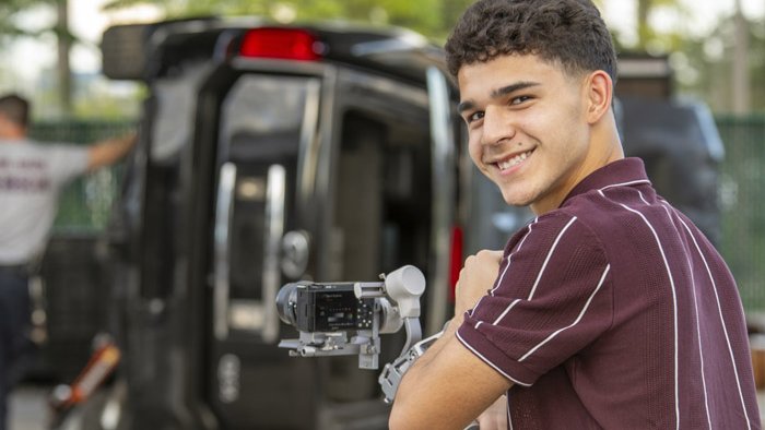 A teenage boy holding his video camera on a gimbal, capturing the scene of a mock accident, while looking back at the camera taking the photo smiling.