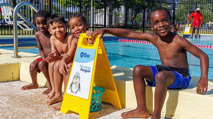 Young children sitting at the edge of a swimming pool, smiling at the camera.